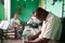 Photo of an older man sitting in a chair in a bright room. His head is bowed and he is folding his hands. He is praying. There are people behind him talking in the room.