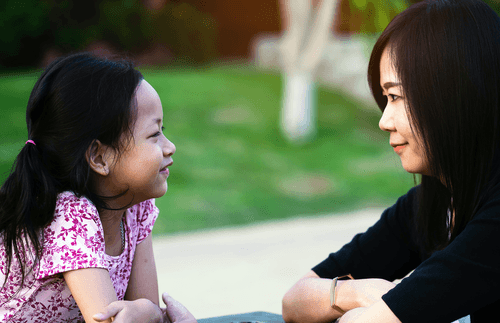 A mother and her daughter are sitting at a picnic table outside, smiling, and looking at each other. The are happy as they share this moment together.